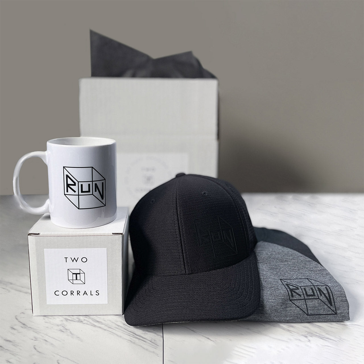 RUN gift box set from TWO CORRALS Lifestyle Running Brand with our signature RUN cube design on a heathered gray and black performance raglan tshirt, white dishwasher safe mug, and black performance hat, wrapped and packaged in our branded gift boxes